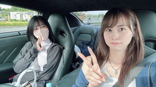 YouTuberあま猫がグレー系スウェットで黒いスポーツカーの助手席からピースサイン。緑豊かな郊外ドライブのセルフィーで、親しみやすい笑顔と車内Vlogの魅力が光る。車好きYouTuberの日常をSNSで発信。