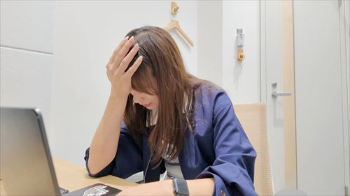 YouTuberあま猫が青いワンピースでデスクワーク中、疲れた表情で頭を抱えるユーモラスな一コマ。シンプルな室内とノートPCが背景のリアルなVlogシーン。SNSで人気のライフスタイルコンテンツ。