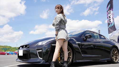 YouTuberあま猫がグレー系フーディーで黒いGT-Rの横で自信たっぷりの後ろ姿。HKSフラッグが背景のサーキットイベントで、車好きYouTuberのSNSで話題のモータースポーツシーン。