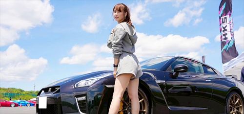 YouTuberあま猫がグレー系フーディーで黒いGT-Rの隣でクールに振り返る。HKSフラッグと青空が背景のサーキットイベントで、車好きYouTuberのダイナミックなSNSショット。